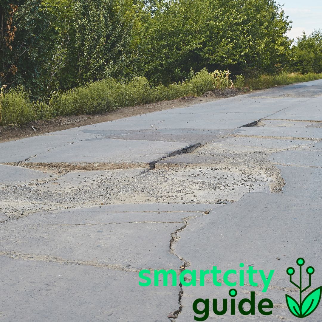 Eine Asphaltstraße mit zahlreichen Schlaglöchern, umgeben von Büschen und Bäumen, mit "smartcity guide"-Logo.