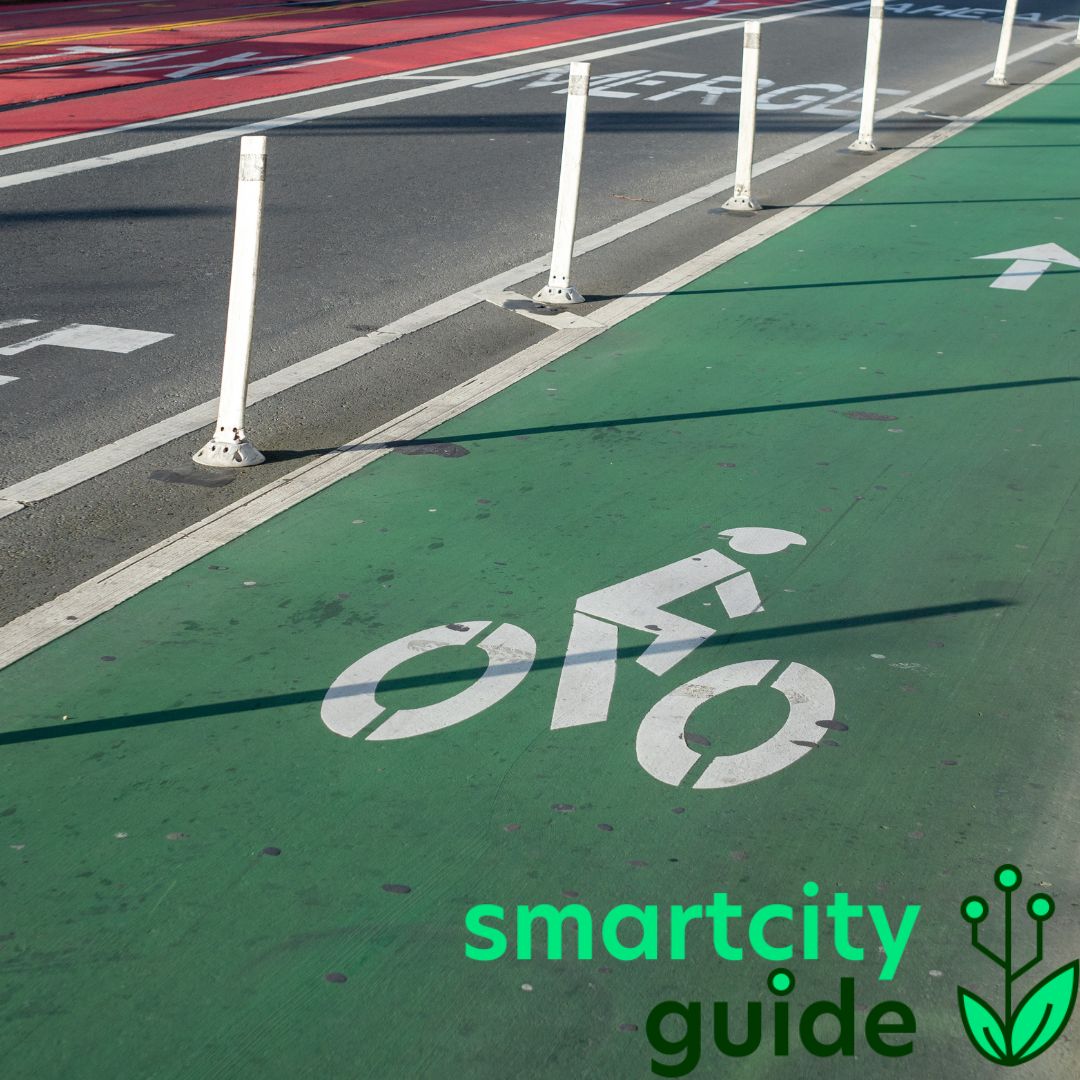 Grüner Fahrradweg mit weißen Markierungen und "smartcity guide" Logo, Stadtstraße im Hintergrund.
