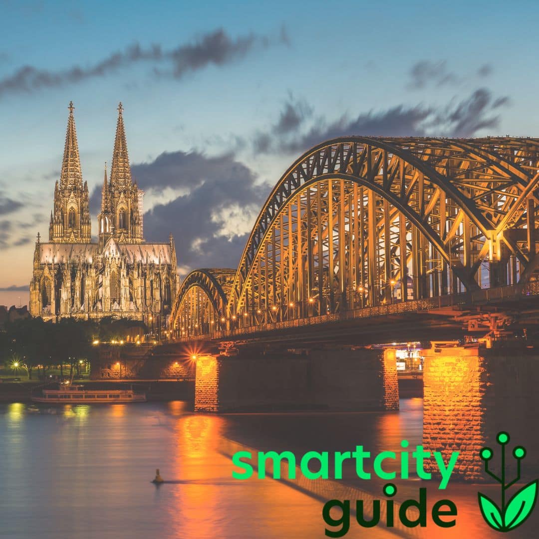 Kölner Dom und Hohenzollernbrücke bei Nacht, beleuchtet und reflektiert im Rhein, mit "smartcity guide"-Logo.